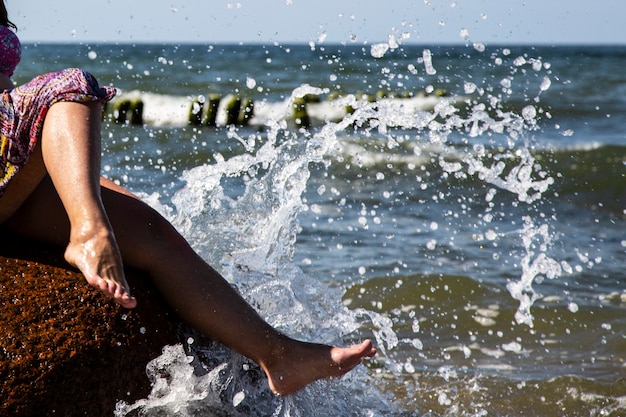 La vacanza in vacanza. Primo piano delle gambe di donna di una ragazza seduta su una grande roccia in riva al mare, onde e spruzzi d'acqua ai suoi piedi in una giornata di sole estivo.