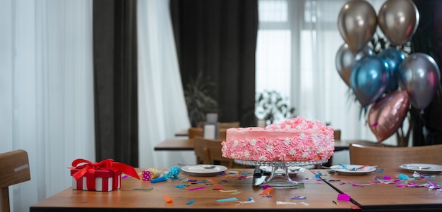 La vacanza per bambini sul tavolo è una torta di compleanno rosa, una scatola rossa con un fiocco e palloncini.