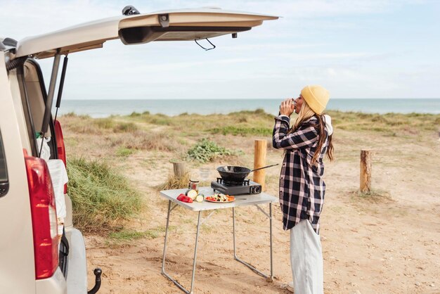 La viaggiatrice prepara il cibo su un fornello a gas portatile su un tavolo pieghevole in un tranquillo punto costiero fuori dal suo camper