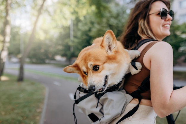 La viaggiatrice sorridente felice sta guidando il suo scooter elettrico nel parco cittadino con il cane Welsh Corgi Pembroke in uno zaino speciale