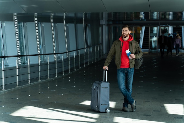 La vista a tutta lunghezza del giovane fiducioso è in piedi nella hall con la valigia e aspetta il volo in aeroporto. Concetto di viaggio