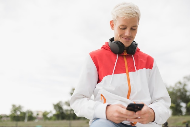 La vista dal basso dell'atleta dell'uomo caucasico si rilassa dopo l'allenamento messaggistica all'aperto tramite i social network su smartphone e utilizzando le cuffie per ascoltare musica Stile di vita sano e concetto di persone