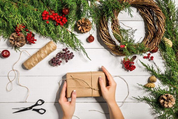 La vista dall'alto delle mani femminili avvolge regali di Capodanno confezionati e pergamene rami di abete e strumenti su...