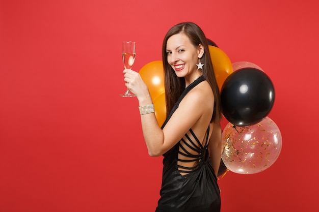 La vista laterale della giovane donna sbalorditiva in vestito nero celebra la tenuta del vetro dei palloni ad aria del champagne isolati su fondo rosso. Concetto di festa di compleanno mockup di San Valentino felice anno nuovo.