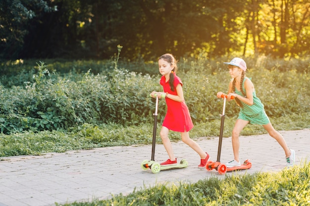 La vista laterale di due ragazze che guidano spinge il motorino su pavimentazione nel parco