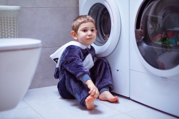 La vista posteriore di un bambino è seduta sul pavimento e guarda nella lavatrice Il bambino in morbido pigiama osserva attentamente il lavaggio