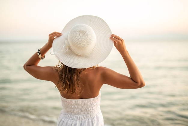 La vista posteriore di una bellissima giovane donna in abito bianco con cappello in testa si rilassa e si gode il tramonto sulla spiaggia.