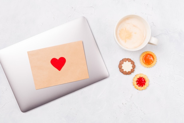 La vista superiore del computer portatile chiuso con la busta del mestiere e cuore rosso, tazza di caffè e vengono biscotti. Desktop.