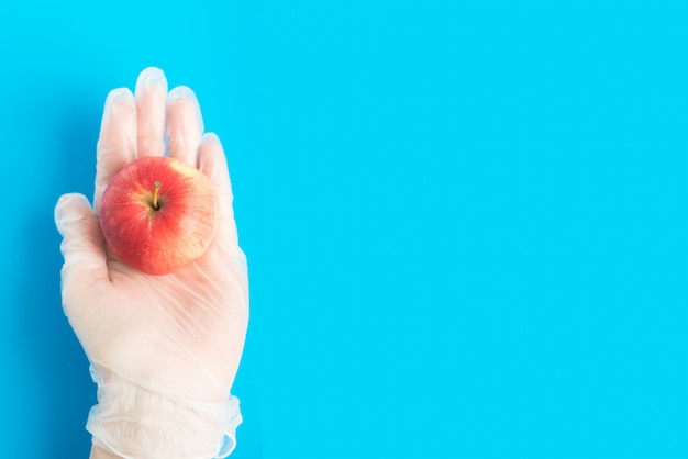 La vista superiore della mano in guanto di gomma tiene la mela rossa sui precedenti blu con copyspace. Concezione di consegna sicura