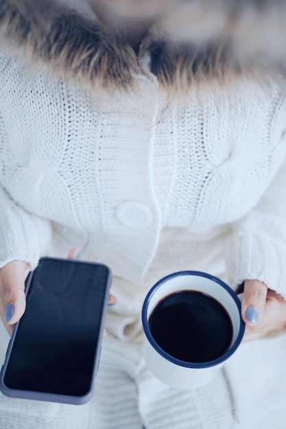 La vista superiore e la fine sulla donna passa la tenuta della tazza di caffè e dello smartphone caldi nella stagione fredda