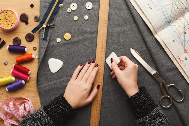 Laboratorio di cucito o stilista al lavoro. Vista dall'alto sulle mani femminili che segnano il quadrato di stoffa con un pezzo di sapone e un righello. Tavolo disordinato con accessori e modelli da sarta, copia spazio