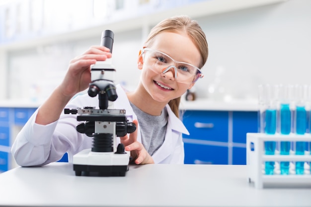 Laboratorio professionale. Ragazza ottimista carina allegra che tocca il microscopio mentre indossa gli occhiali di sicurezza e che guarda l'obbiettivo