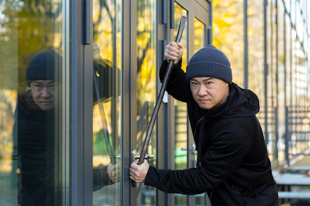 Ladro asiatico cerca di rompere la serratura della porta con un grande strumento di piede di porco di metallo