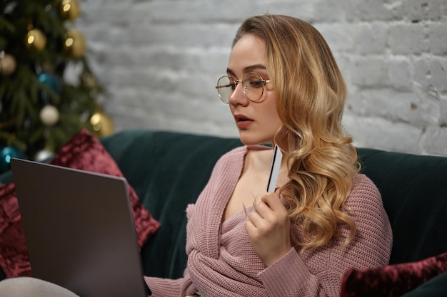 Lady blogger in cardigan rosa pantaloni occhiali seduto sul divano verde con cuscini colorati che mostrano pla...