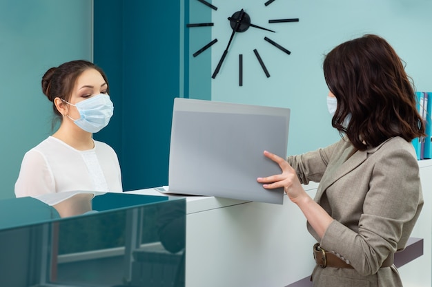 Lady office manager con maschera protettiva mostra il libro di registrazione al cliente donna in piedi alla reception nella vista laterale vicina dell'ospedale contemporaneo