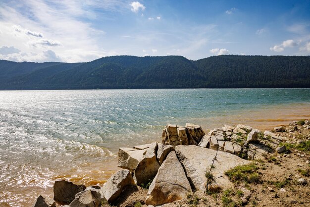 Lago con costa rocciosa sullo sfondo della catena montuosa ricoperta di foresta e cielo blu