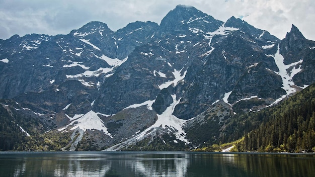 Lago di montagna con acqua blu fredda, foresta alpina e roccia innevata