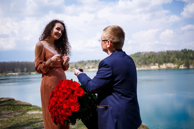 Lago nella foresta: un uomo ha fatto una sorpresa alla sua ragazza, le mette un anello al dito, una dichiarazione d'amore e un'offerta di matrimonio. Ragazza con un sorriso sul viso e un mazzo di rose rosse