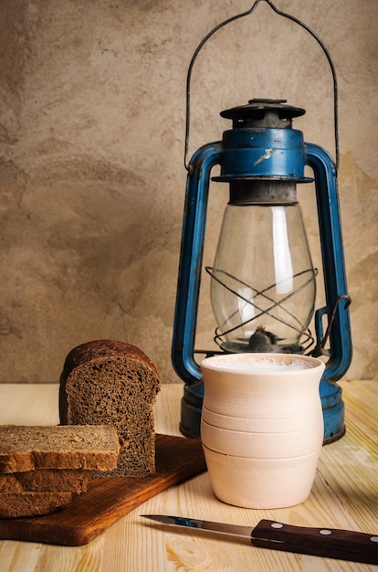 Lanterna di cherosene tagliere vaso di argilla con pane di segale al latte e un coltello su un tavolo di legno