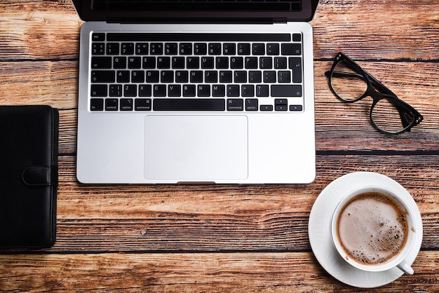 laptop, occhiali, blocco note e una tazza di caffè su una vista dall'alto di un tavolo in legno. Vacanze o concetto di acquisto online