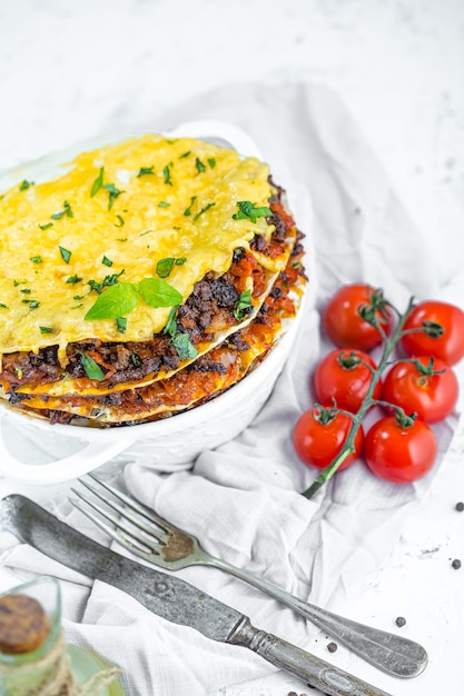 lasagne in una pentola di argilla rotonda bianca profonda, su uno sfondo di marmo bianco, forchetta e coltello giacciono su un panno,