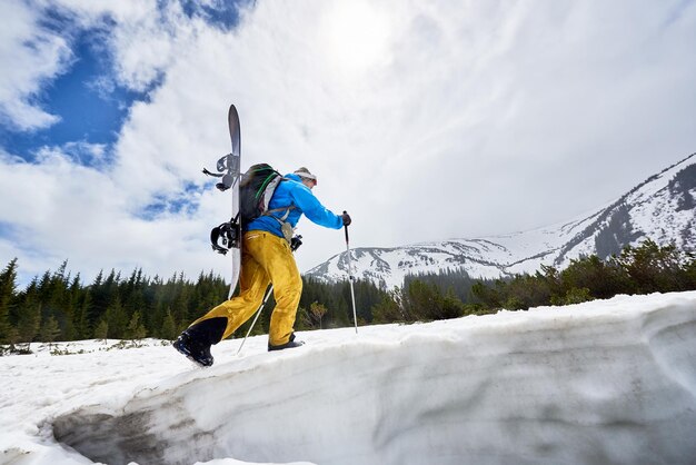 Lato della faccia nascosta dello snowboarder con lo snowboard sulla schiena sul pendio innevato Nuvole sul cielo blu sullo sfondo Angolo basso