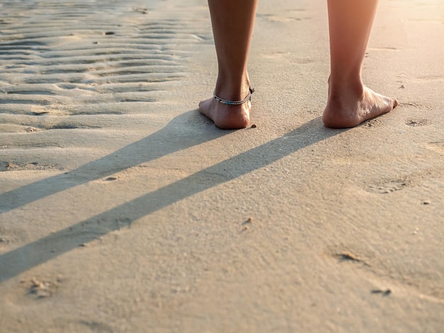 Lato posteriore della donna a piedi nudi in pantaloni bianchi con gambe piegate in piedi sulla spiaggia di sabbia pulita alla calda luce del sole al tramonto con spazio di copia Destinazione di viaggio estiva e concetti di vacanza