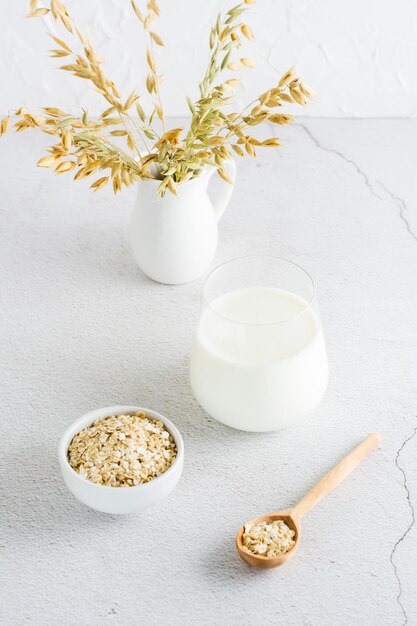 Latte d'avena in un bicchiere di farina d'avena in una ciotola e orecchie in una brocca su un tavolo luminoso Alimenti vegetali alternativi Vista verticale