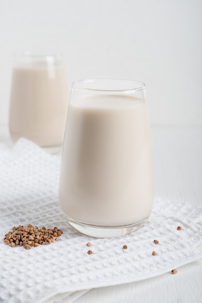 Latte di grano saraceno vegano senza lattosio nel bicchiere con grani crudi sul tovagliolo sul tavolo di legno bianco