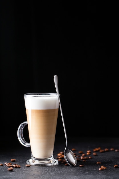 latte macchiato in una tazza di vetro con manico con caffè intorno e cucchiaio isolato su sfondo scuro