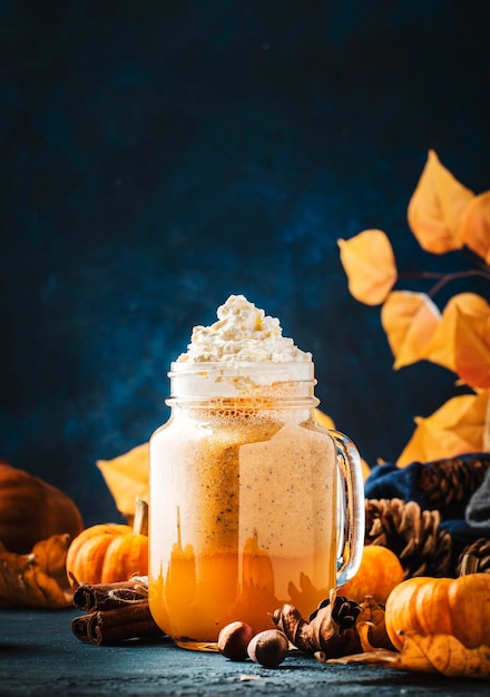 Latte o caffè speziato alla zucca in un vasetto di vetro sul tavolo blu Bevanda calda autunnale o invernale in una tavola festosa naturale con foglie d'arancio spezie piccole pigne di zucca