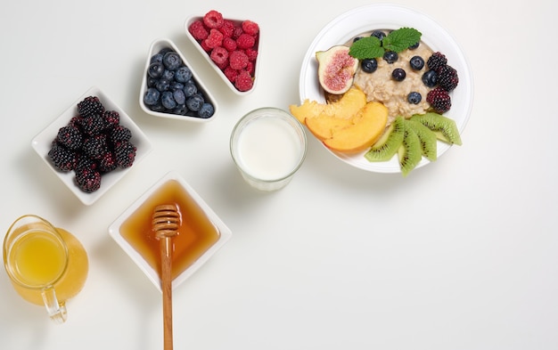 Latte, piatto con porridge di farina d'avena e frutta, succo appena spremuto in un decanter di vetro trasparente, miele in una ciotola su un tavolo bianco. Colazione salutare