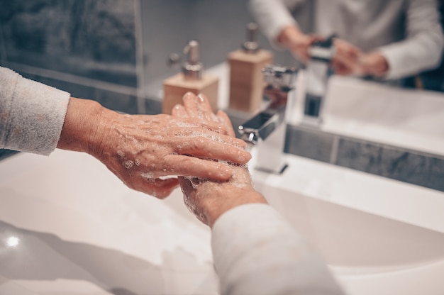 Lavaggio a mano schiuma di sapone liquido sfregamento polsi lavare a mano donna senior passo risciacquo in acqua al lavandino del rubinetto del bagno. Lavarsi le mani per prevenire la diffusione del COVID-19. Focolaio di pandemia di coronavirus.
