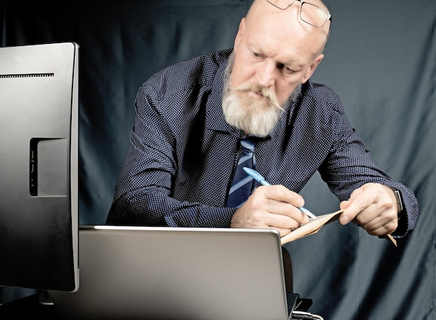 Lavora in ufficio un uomo anziano serio con una barba grigia davanti a un computer portatile scrive su un quaderno