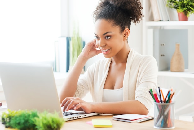 Lavorando al nuovo progetto creativo. Bella giovane donna africana che lavora al computer portatile e sorride mentre è seduta al suo posto di lavoro in ufficio