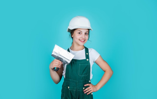 Lavorando nel concetto di officina di riparazione di costruzione e ristrutturazione ragazza felice usa lo strumento spatola costruttore adolescente in casco di sicurezza giovane lavoratore in elmetto protettivo bambino fiducioso usa la spatola durante la riparazione