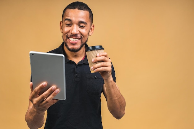 Lavorando su tavoletta digitale Ritratto di felice sorridente giovane nero africano americano azienda tavoletta digitale mentre in piedi isolato su sfondo beige Bere caffè