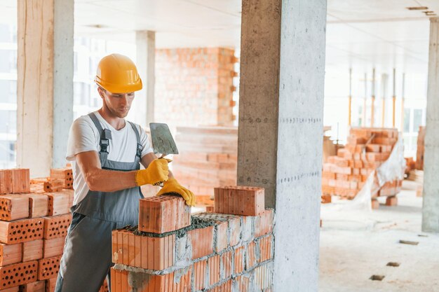 Lavorando usando i mattoni Giovane in uniforme alla costruzione durante il giorno