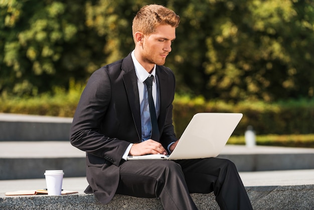 Lavorare all'aperto. Fiducioso giovane in camicia e cravatta che lavora al computer portatile mentre è seduto all'aperto