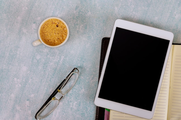 Lavorare alla scrivania in ufficio utilizzando una tavoletta digitale con tazza di porcellana di caffè nero, occhiali da vista, notebook settimanale