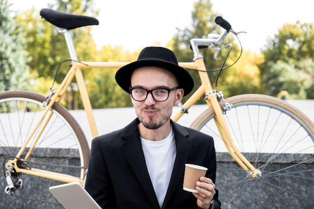 Lavorare con la tecnologia fuori dal concetto di ufficio. Ritratto di giovane con l'espressione del viso allegro divertente in abiti casual alla moda che si siede con computer tablet e caffè all'aperto