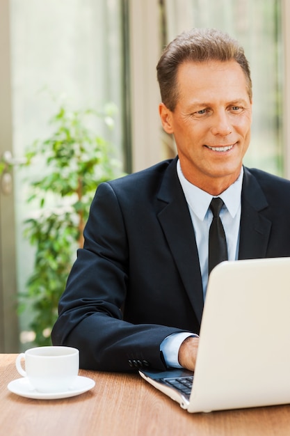 Lavorare con piacere. Uomo maturo allegro in abiti da cerimonia che lavora al computer portatile e sorride mentre è seduto al tavolo all'aperto