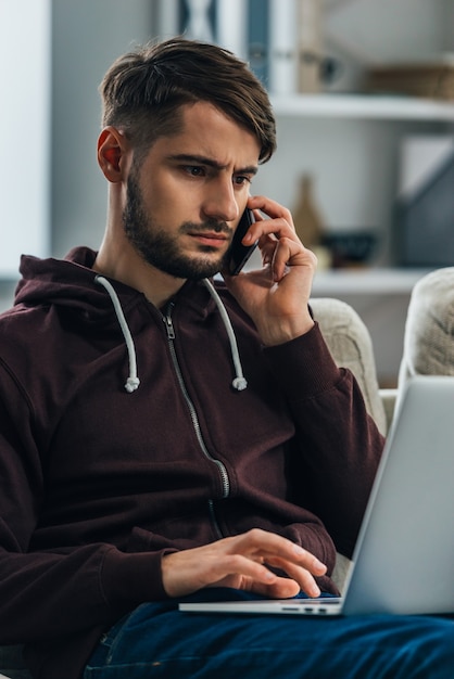 Lavorare da casa. Giovane concentrato che usa il suo laptop e parla al cellulare mentre è seduto sul divano di casa