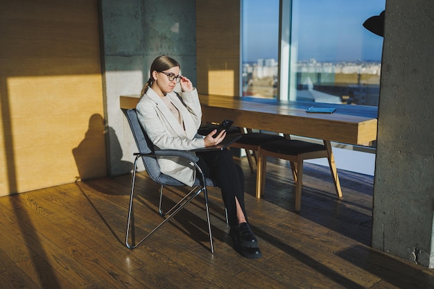 Lavorare in un ufficio moderno Positiva giovane donna d'affari in abiti casual e occhiali seduti in ufficio con un telefono cellulare mentre si lavora su un progetto remoto utilizzando un computer portatile
