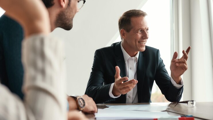 Lavorare insieme Uomo maturo bello e felice in abiti formali che gesticola e sorride mentre è seduto con i suoi colleghi in ufficio