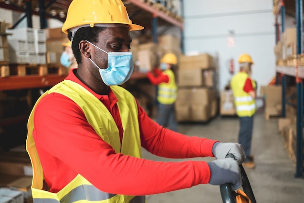 Lavoratore afroamericano all'interno del magazzino che tira un transpallet mentre indossa la maschera di sicurezza