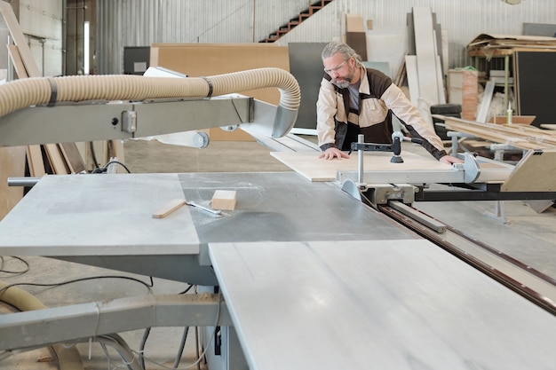 Lavoratore anziano di mobili che produce in fabbrica il fissaggio di una tavola rettangolare sul banco da lavoro prima di tagliarla o molarla con un utensile elettrico