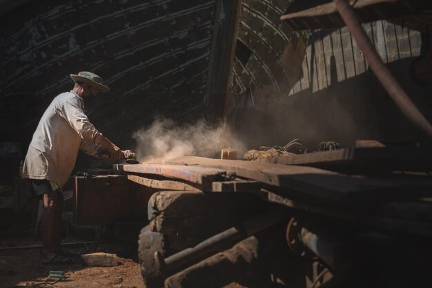 Lavoratore asiatico locale che macina legno vecchio in cantiere navale per la manutenzioneSituazione reale della fabbrica locale asiaticaconcetto di vita del lavoratore industriale