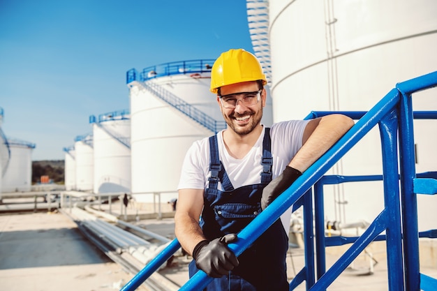 Lavoratore caucasico bello sorridente nel complesso e con il casco sulla testa che si appoggia l'inferriata. Produzione di olio.