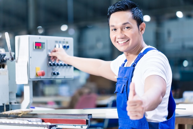 Lavoratore che mostra i pollici in su in un impianto di produzione asiatico in piedi davanti a un pannello di controllo della macchina